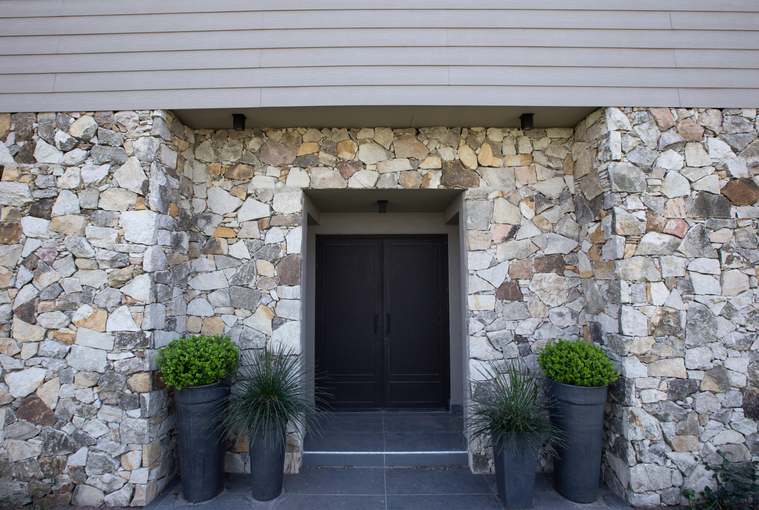 Modern architecture. Residential facade design. Closeup view of new house iron front door, stonewall and decorative plant Buxus sempervirens, also known as boxwood bush, growing in pots.